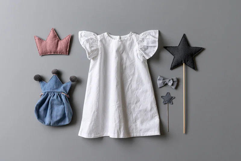 White cotton toddler dress with flutter sleeves arranged flat on gray surface with felt crowns, star wand and bowtie accessories