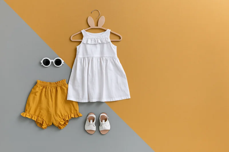 Baby girl summer outfit flat lay with white dress, yellow shorts, sandals and sunglasses on two-tone background
