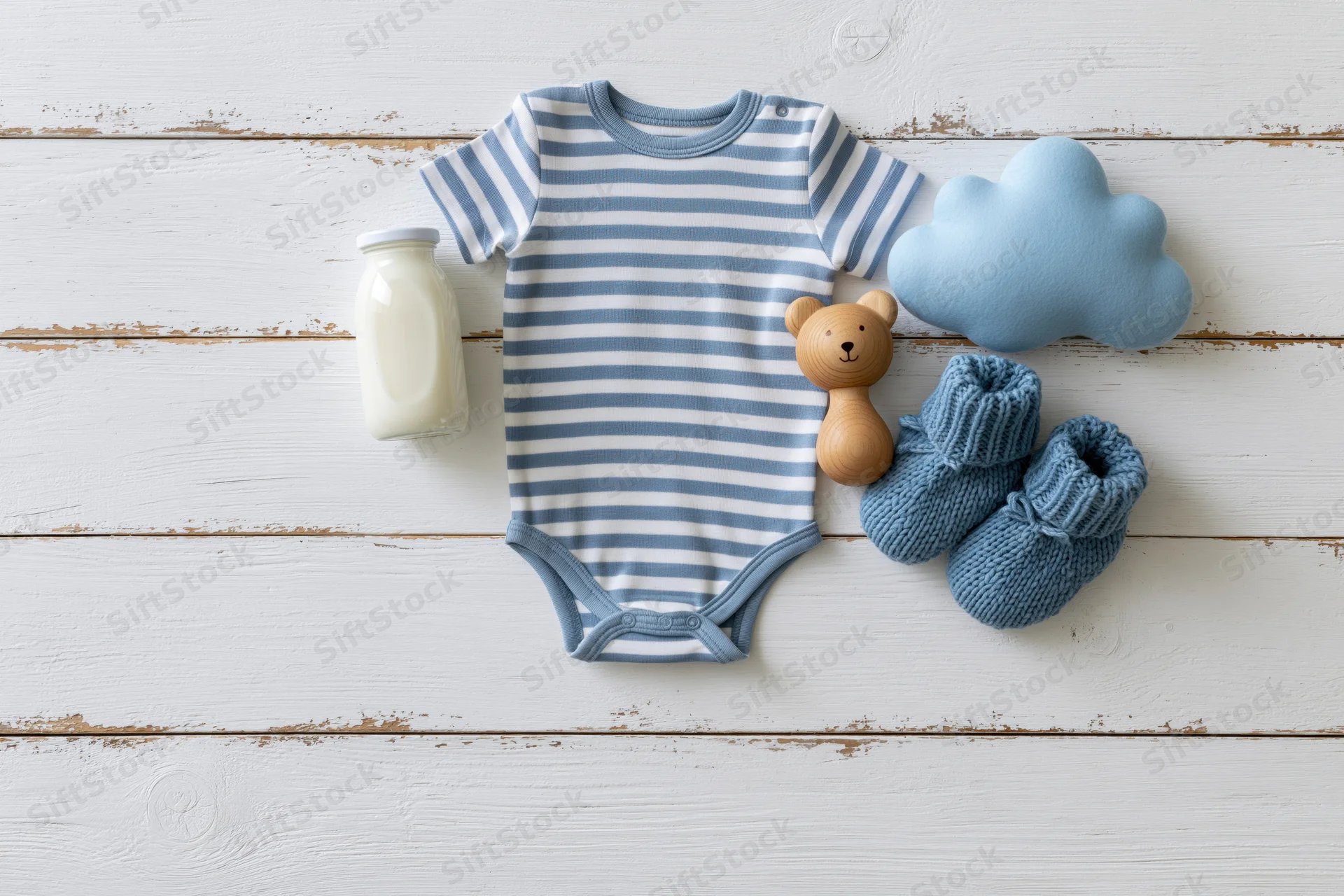 Baby onesie with knitted booties, wooden bear toy, milk bottle and cloud plush arranged on white wooden planks