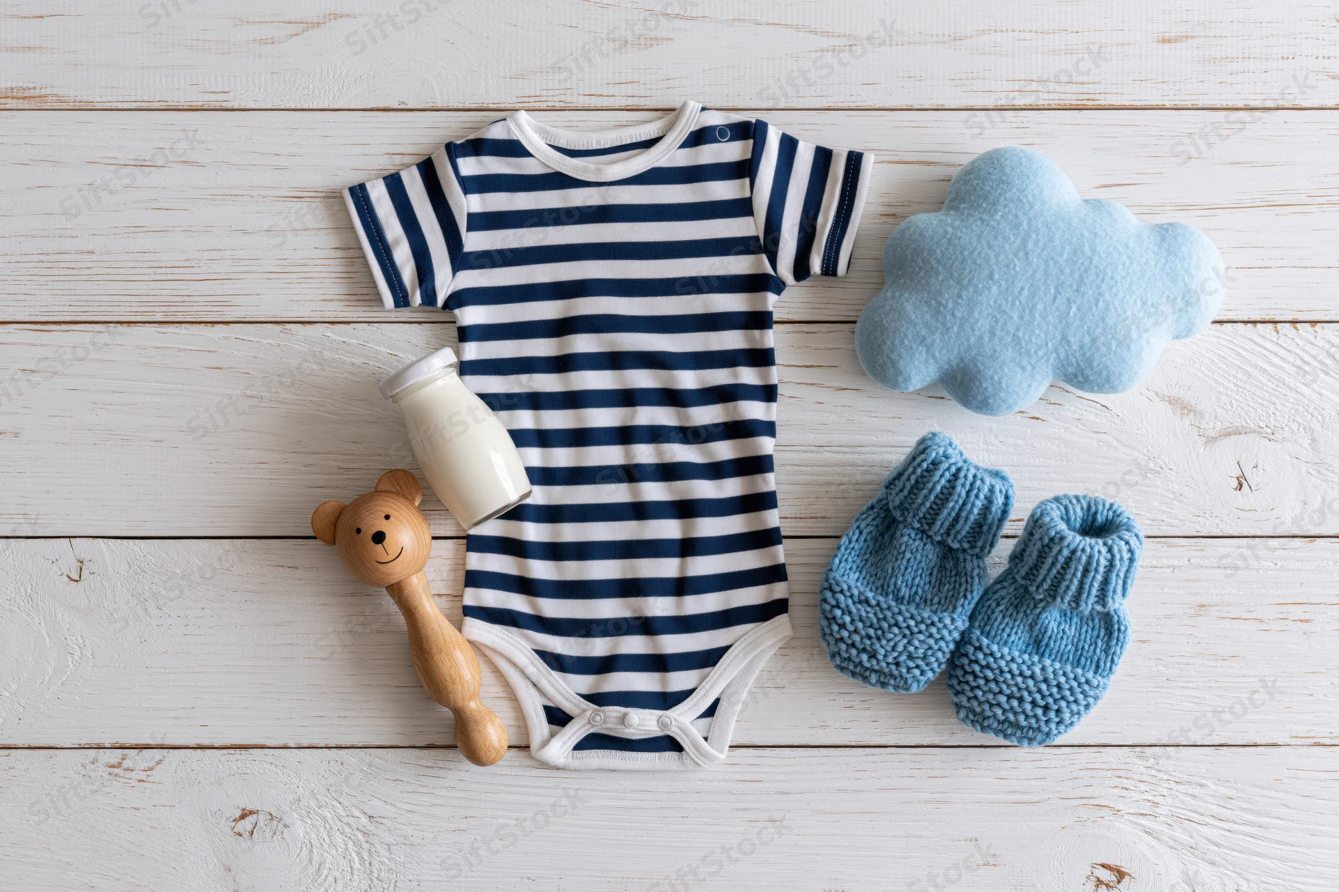Baby onesie with striped pattern, knitted booties, wooden rattle, milk bottle and felt cloud arranged on white painted wooden plank