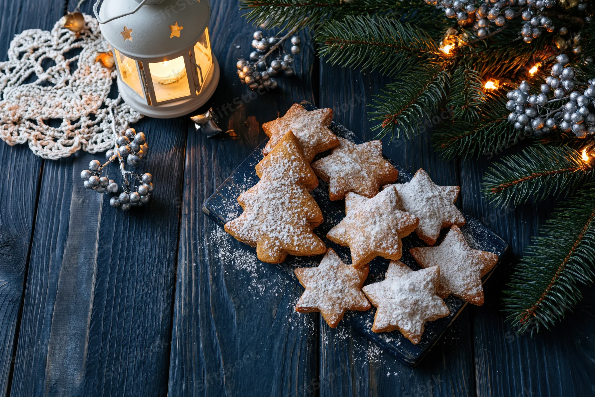 Star and tree shaped gingerbread cookies dusted with sugar on a dark wooden table surrounded by pine branches, silver ornaments, and a glowing lantern
