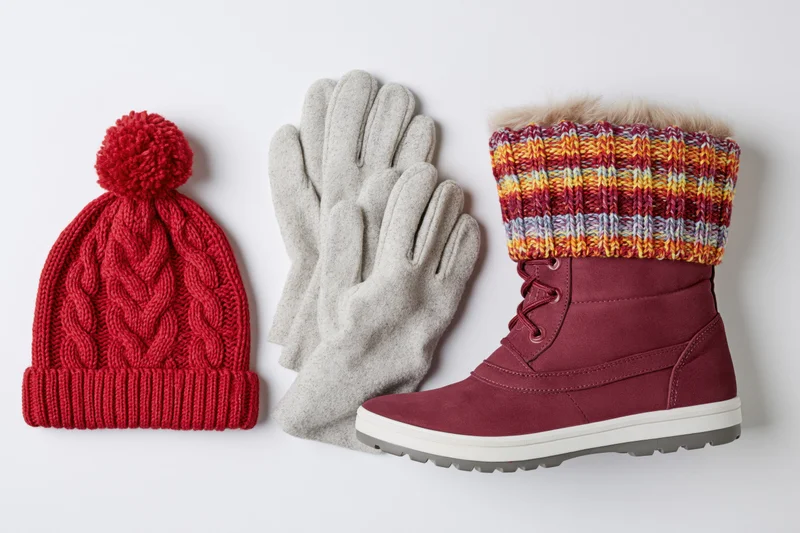 Winter accessories including a red knit hat, gray gloves, and a maroon boot with striped knit cuff arranged on a white surface
