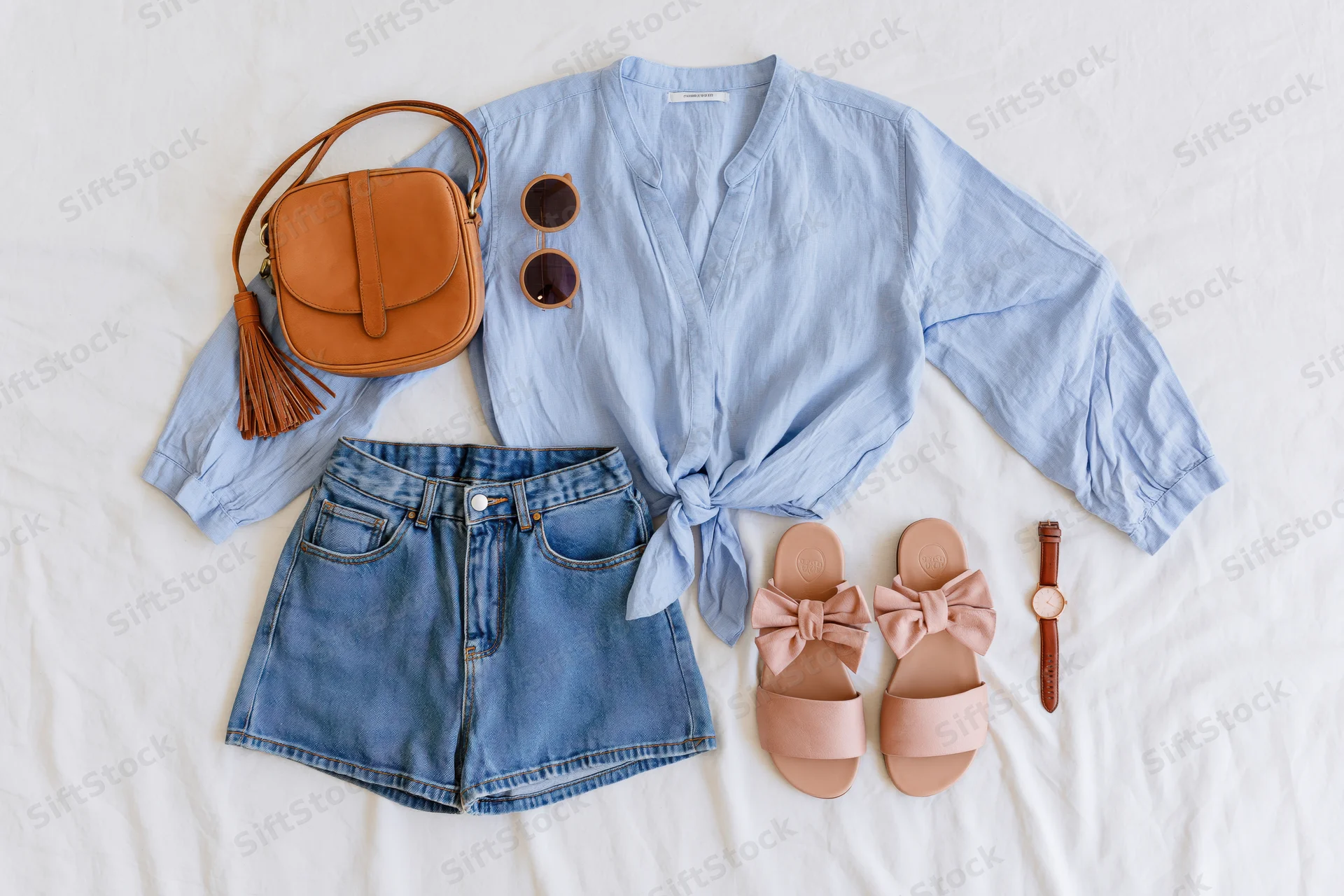 Women's summer outfit flat lay with denim shorts, blue blouse, sandals, bag and accessories on white cotton sheet