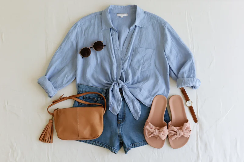 Women's summer outfit with denim shorts, blue shirt tied at waist, sunglasses, leather bag, pink bow sandals and watch arranged on white cotton fabric