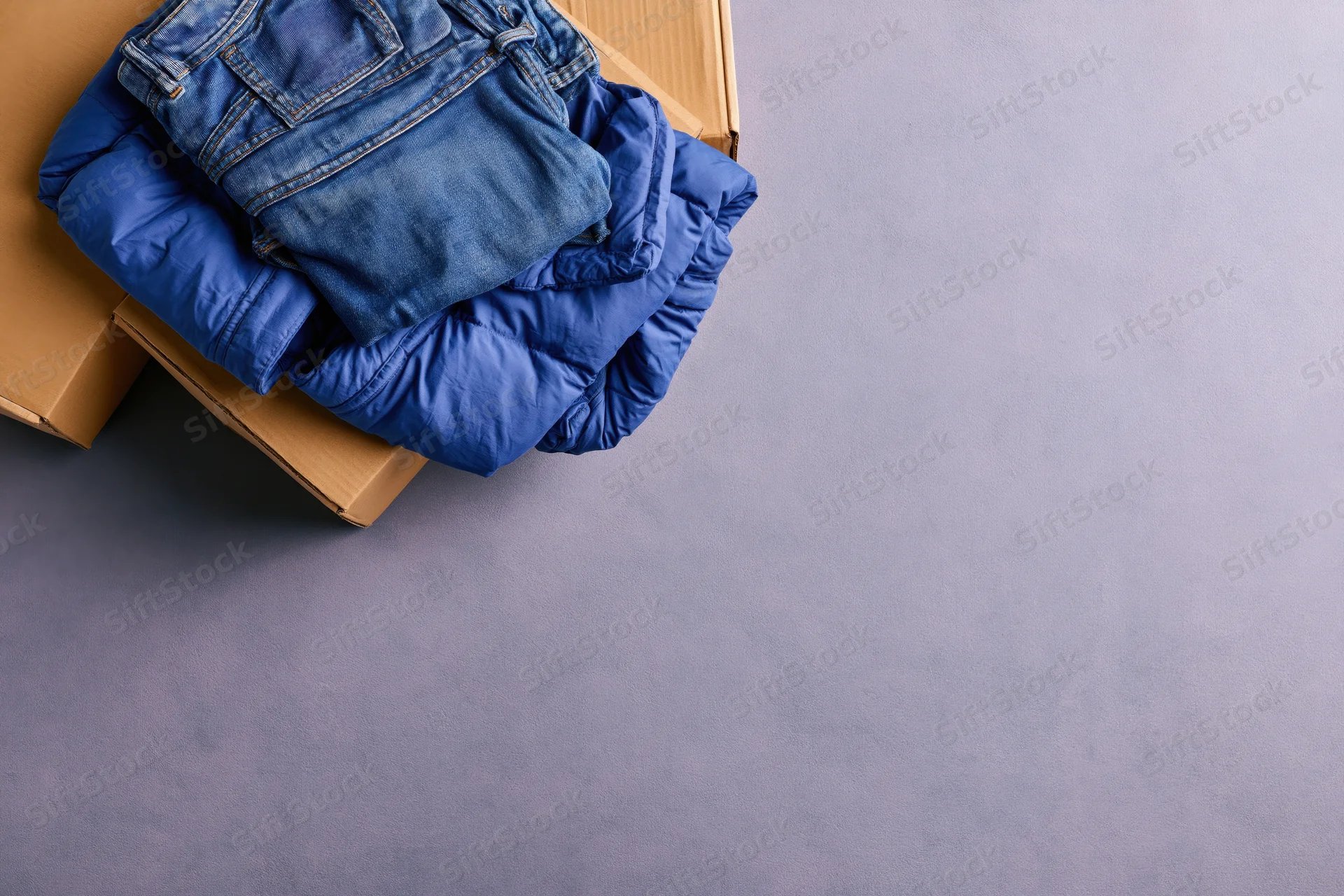 Folded blue denim jeans and royal blue puffer jacket stacked on cardboard box on gray surface