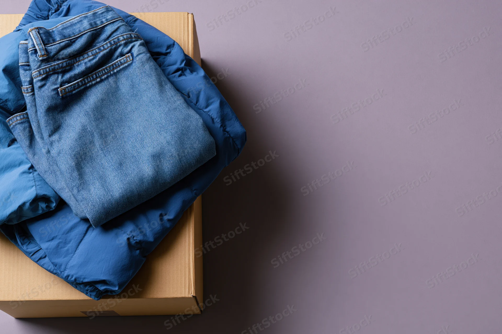 Folded blue denim jeans and royal blue puffer jacket stacked on cardboard box on gray surface