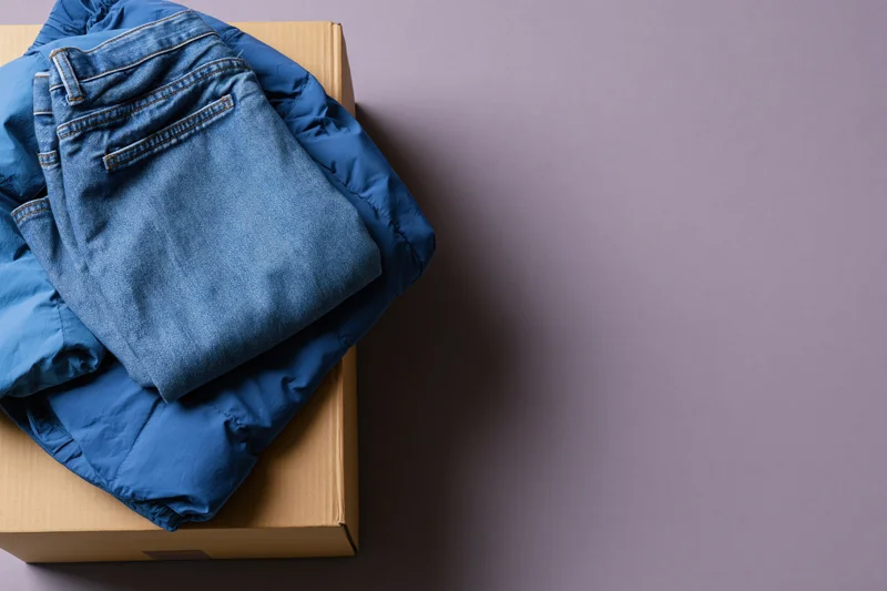 Folded blue denim jeans and royal blue puffer jacket stacked on cardboard box on gray surface
