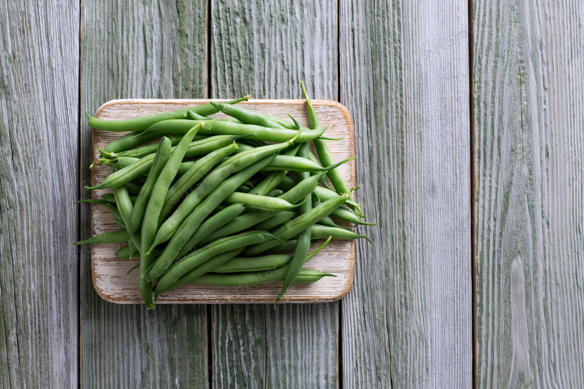Fresh green beans piled on a small worn wooden cutting board on a rustic wooden table