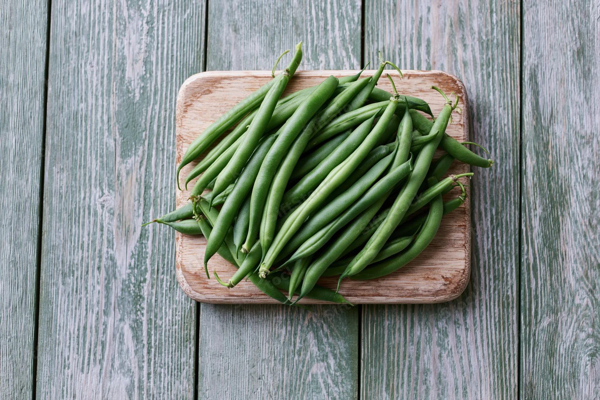 Fresh green beans piled on a wooden cutting board on a weathered wood surface