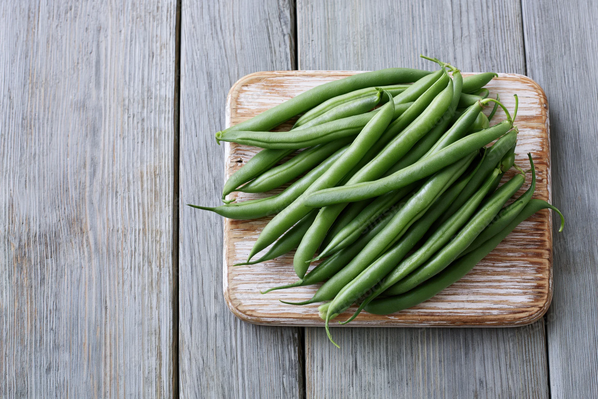 Fresh green beans piled loosely on a small worn wooden cutting board on a gray weathered wooden surface