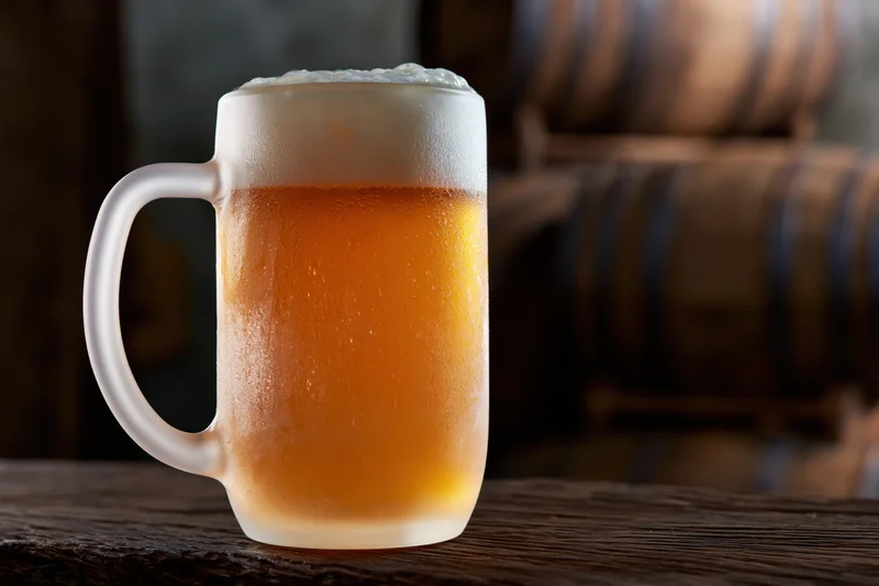 Frosted glass beer mug filled with pale lager and thick foam on wooden table with barrels in background