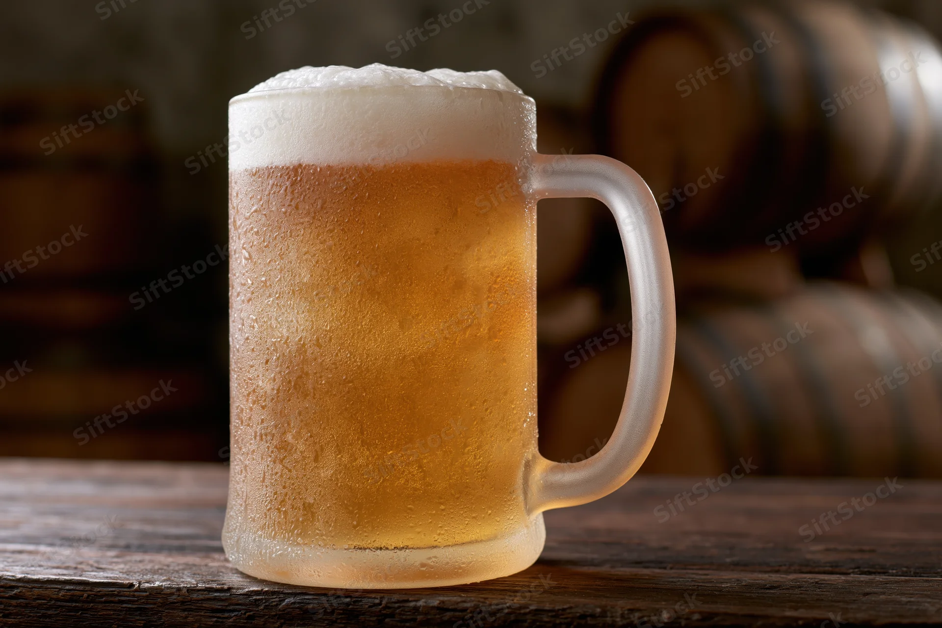 Frosted glass beer mug filled with pale lager and thick foam on wooden table with barrels in background