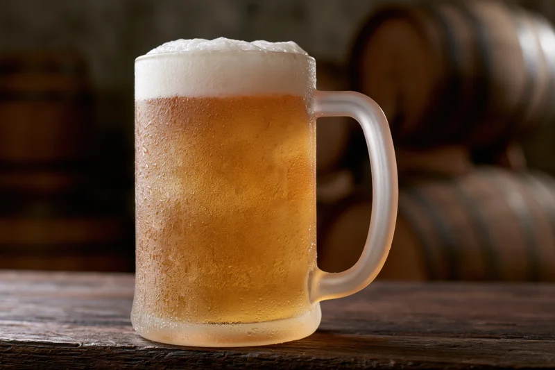 Frosted glass beer mug filled with pale lager and thick foam on wooden table with barrels in background