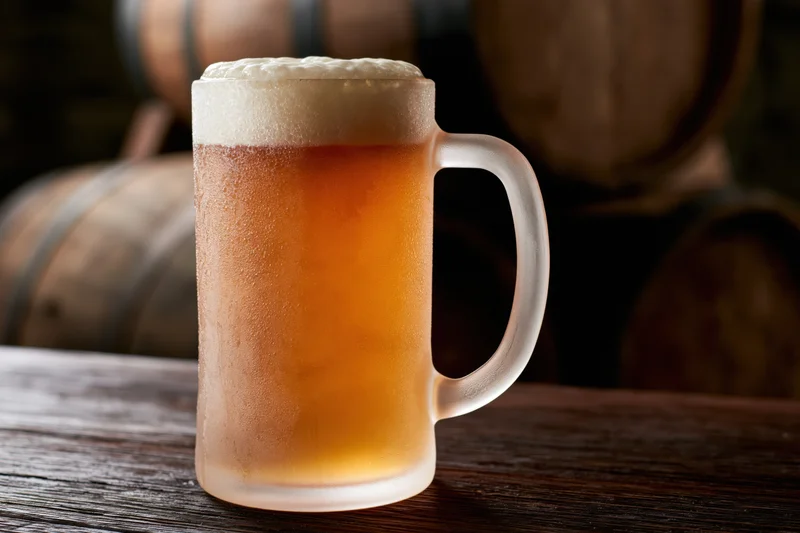 Frosted glass beer mug filled with pale lager and thick foam on wooden table with barrels in background