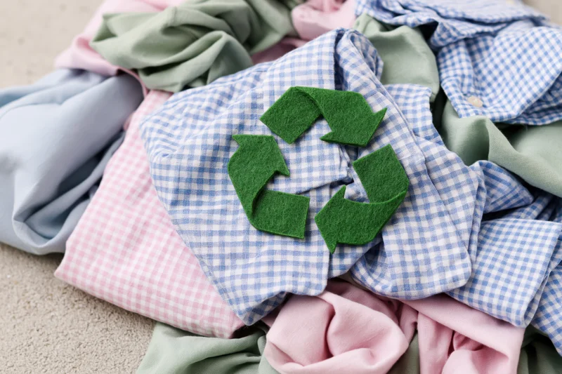 Green felt recycling symbol on a pile of mixed clothing including checkered shirts and pastel fabrics