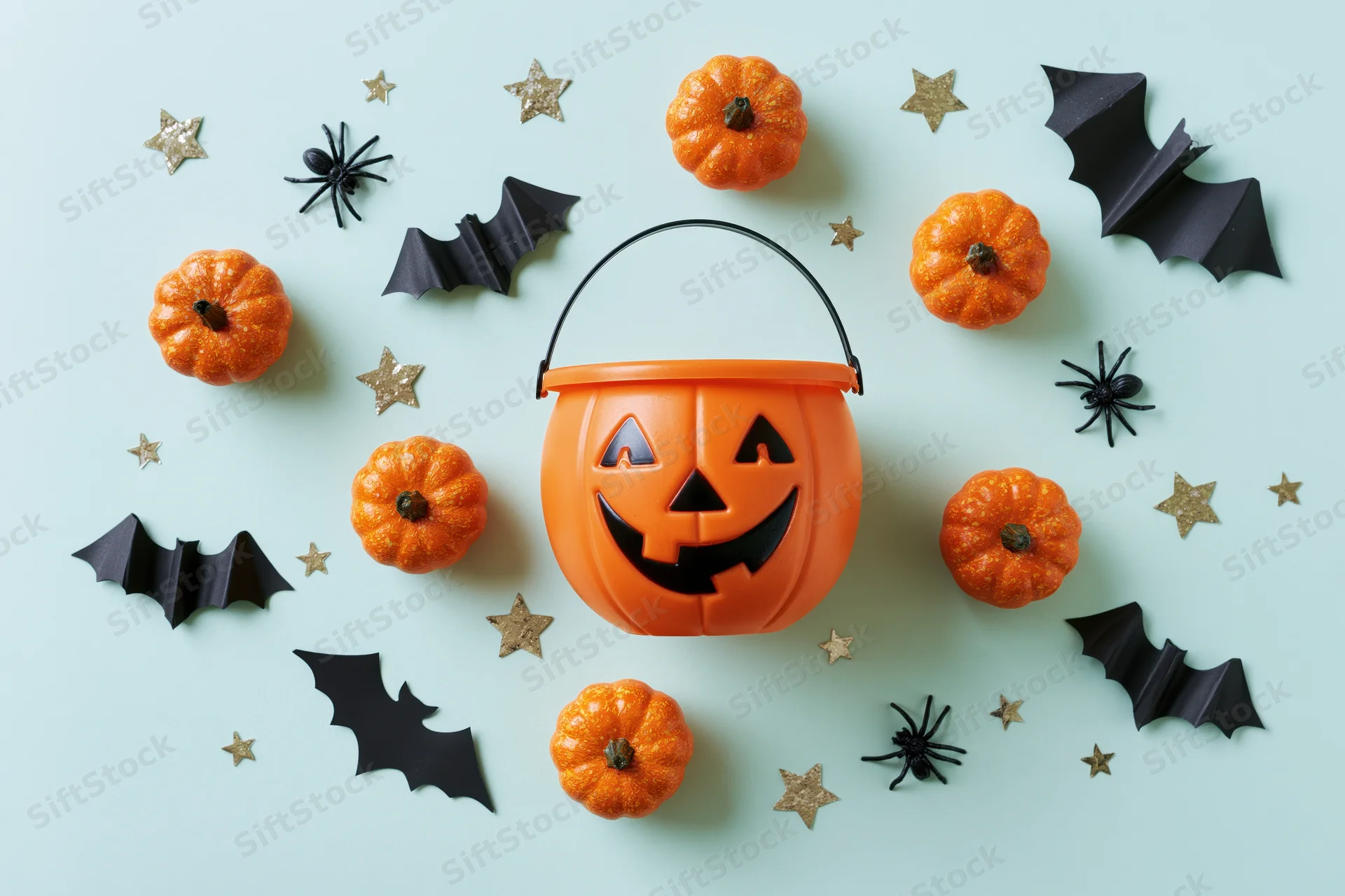 Orange jack-o-lantern candy bucket surrounded by small pumpkins, black bats, spiders and glitter stars on mint background