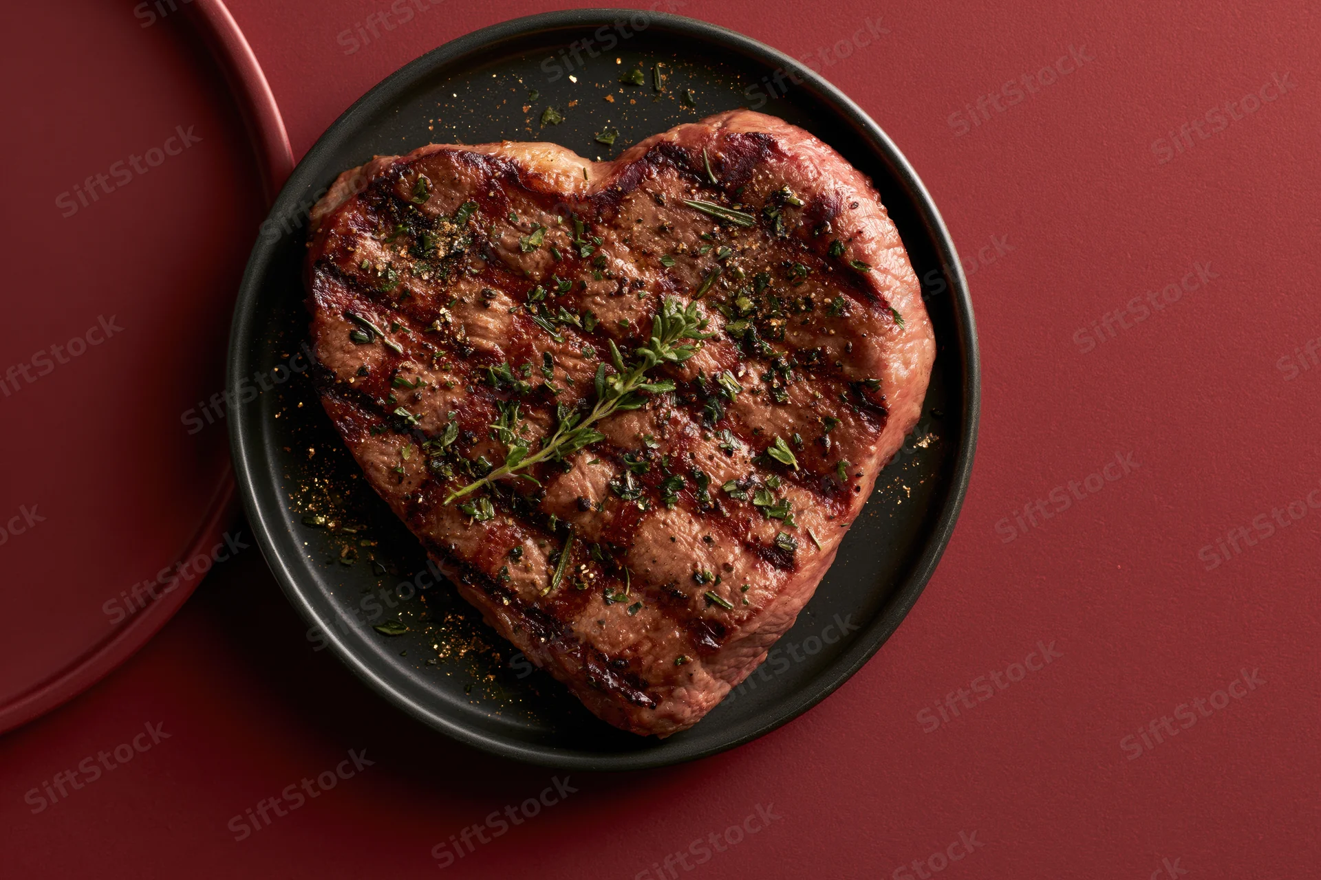 Heart-shaped grilled steak topped with fresh herbs on a black plate against a red background