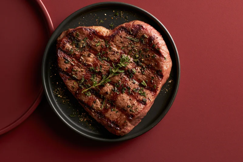 Heart-shaped grilled steak topped with fresh herbs on a black plate against a red background