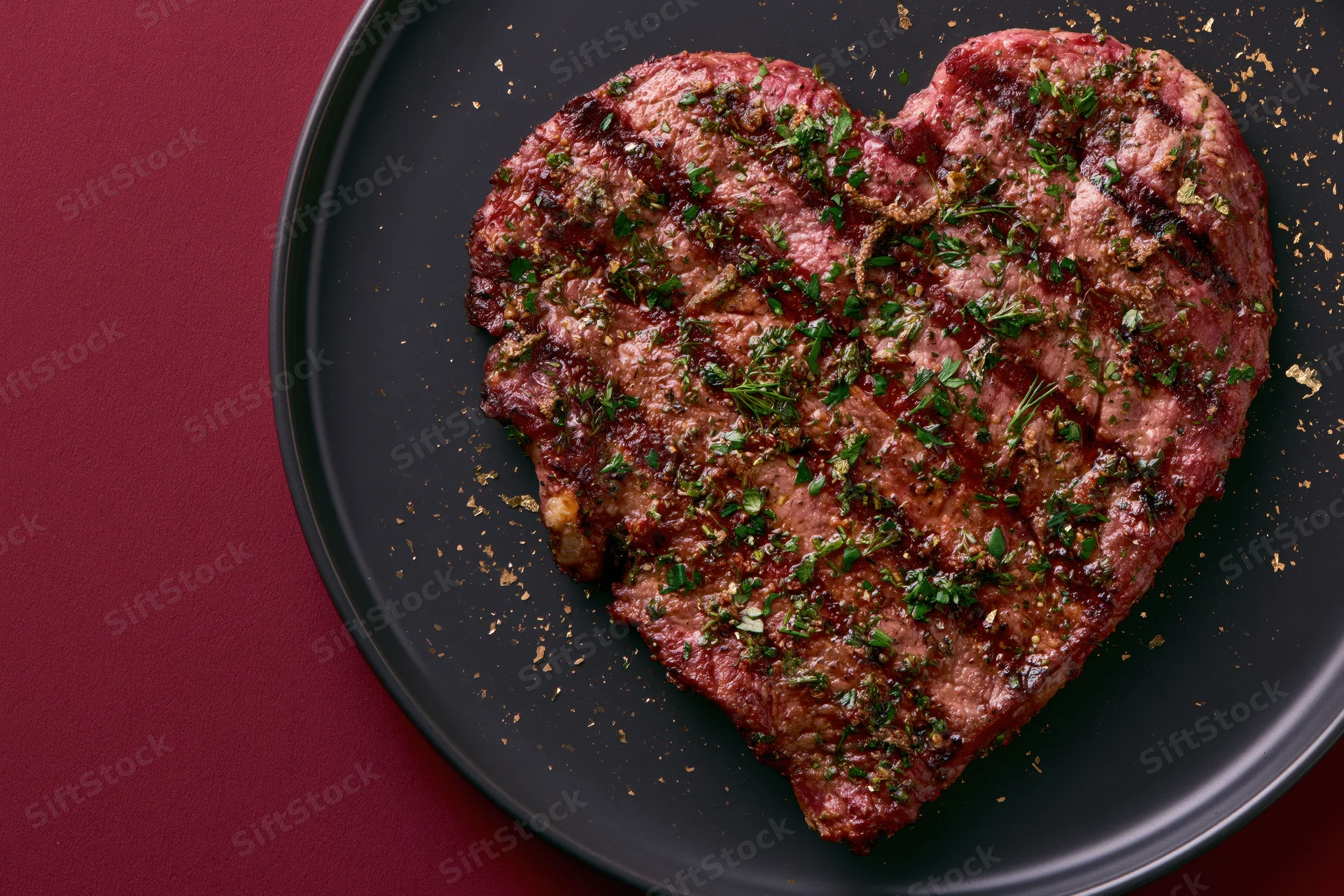 Heart-shaped grilled steak topped with fresh herbs on a black plate against a red background