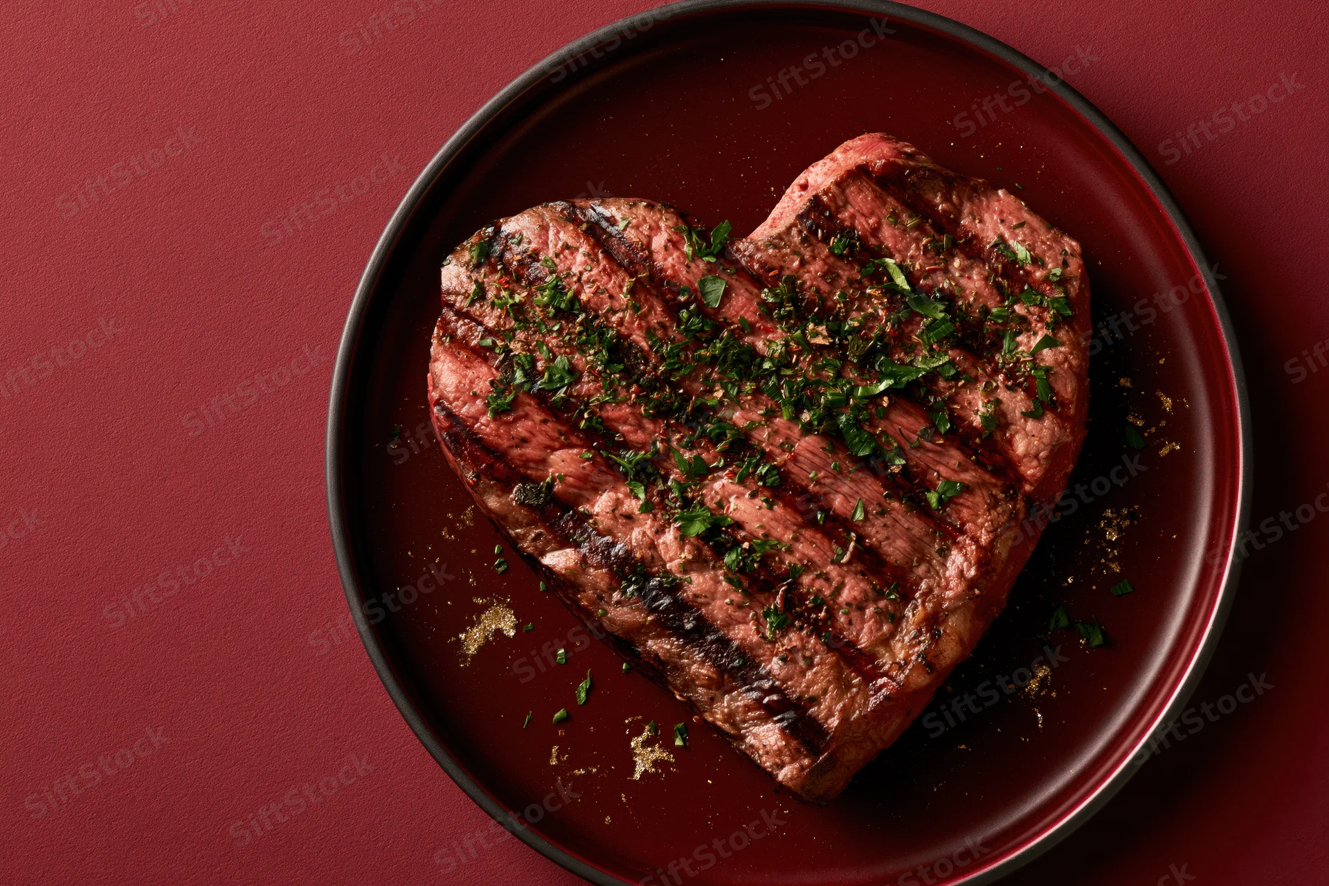 Heart-shaped grilled steak topped with herbs on a dark red plate