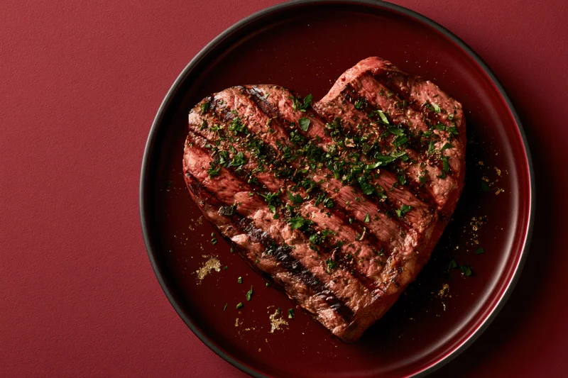 Heart-shaped grilled steak topped with herbs on a dark red plate