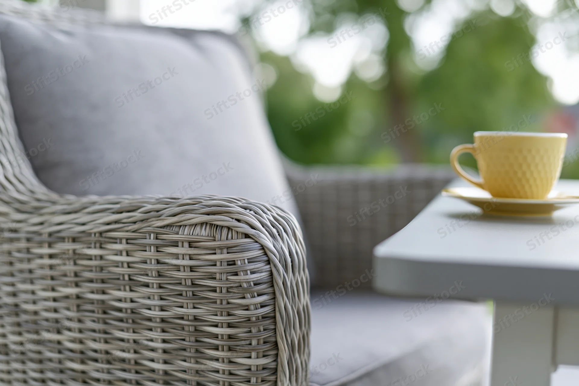 Light grey woven rattan outdoor armchair with soft cushion beside a small table holding a yellow coffee cup and saucer, set on a patio with green foliage in the background