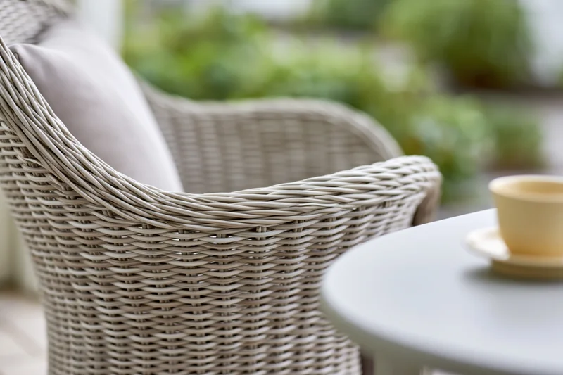 Light grey woven rattan outdoor armchair with soft cushion beside a small table with a cup and saucer in a garden setting