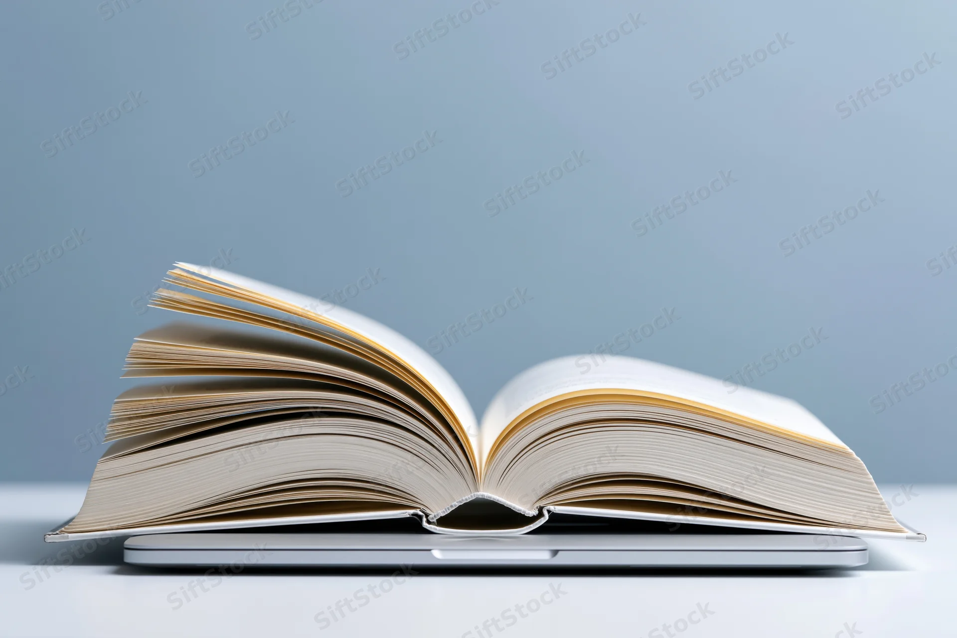 Open hardcover book with fanned pages resting on a closed laptop on a plain surface