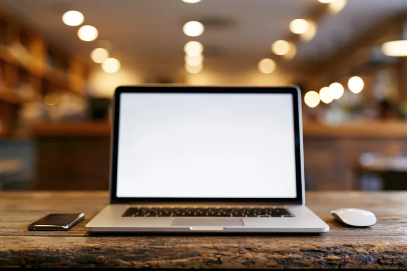 Open laptop with blank white screen on wooden table in cafe interior