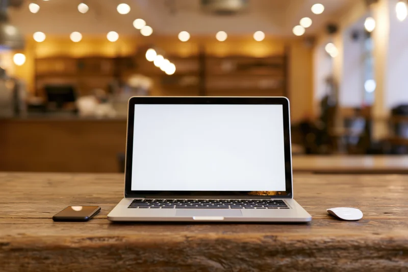 Open laptop with blank white screen on wooden table in cafe interior