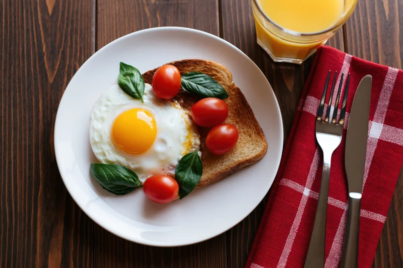 Fried egg on toast with cherry tomatoes and basil on a white plate, accompanied by orange juice and cutlery on a wooden table