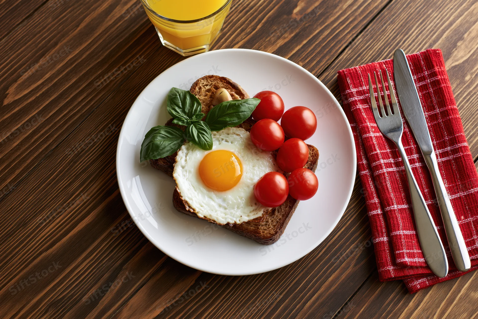 Fried egg on toast with cherry tomatoes and basil on a white plate with orange juice and cutlery on a wooden table
