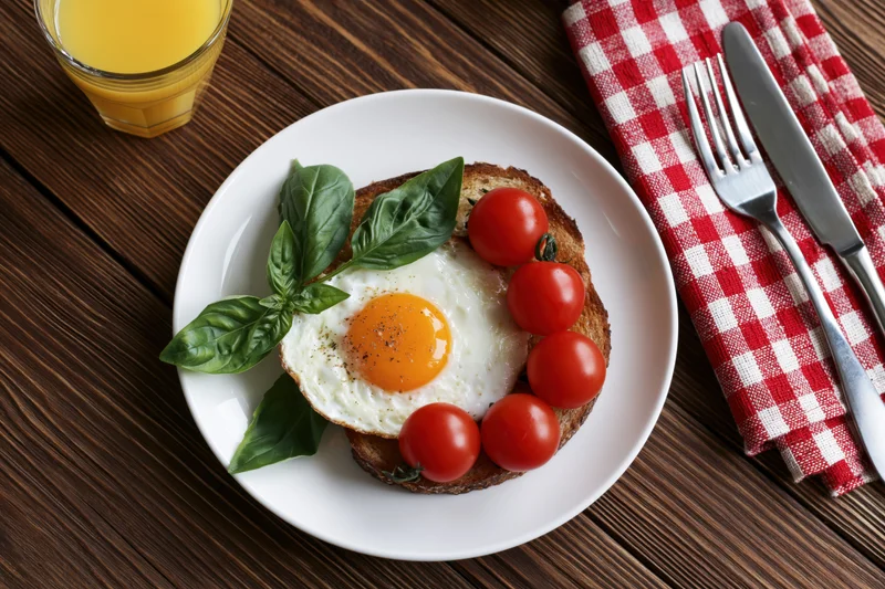 Fried egg on toast with cherry tomatoes and basil on a white plate, served with orange juice and cutlery on a wooden table with a red checkered napkin