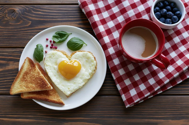Heart-shaped fried egg with toast and coffee on a dark wood table with checkered napkin
