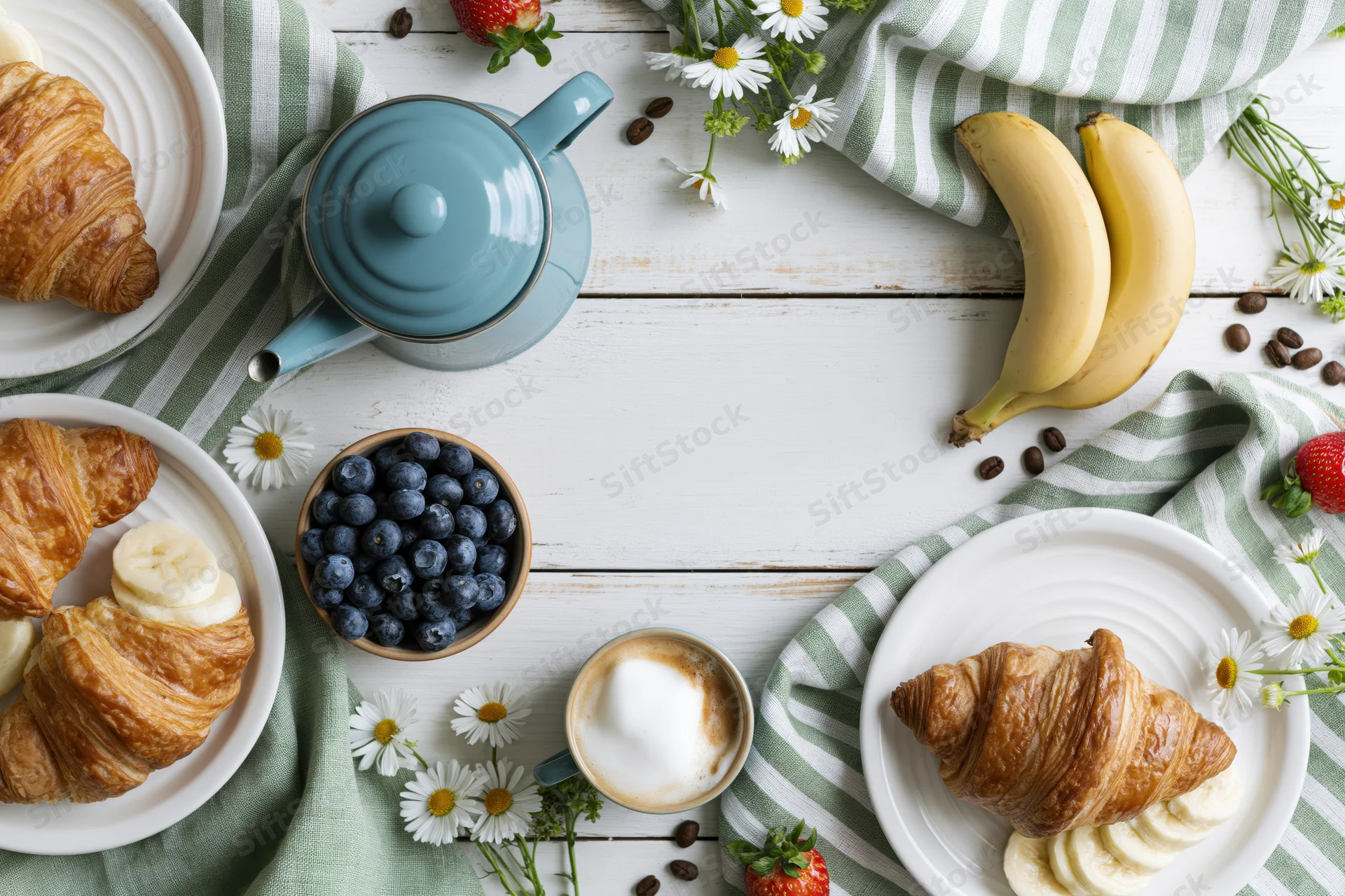 Overhead flat lay of breakfast items including croissants, bananas, blueberries, coffee, and daisies on a white wooden table with striped napkins