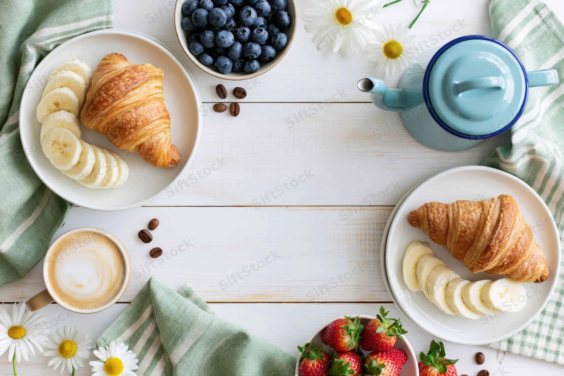 Overhead flat lay of breakfast with croissants, bananas, berries, coffee and daisies on white wooden table