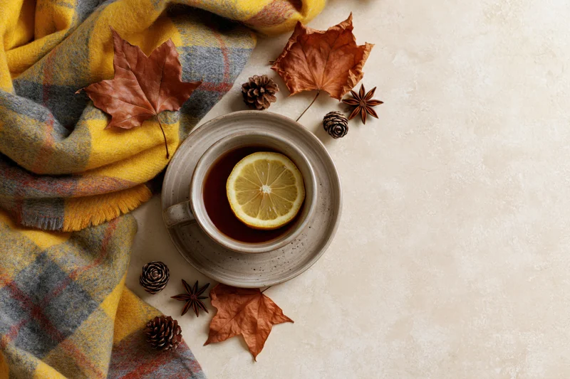 Ceramic cup of tea with lemon slice on a flat lay with autumn leaves, pinecones, and a plaid scarf