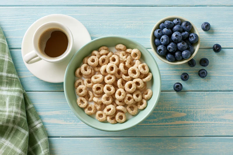 Overhead flat lay of a green bowl with cereal and milk, blueberries in a small bowl, coffee cup on saucer, on a blue wooden table with a green plaid cloth