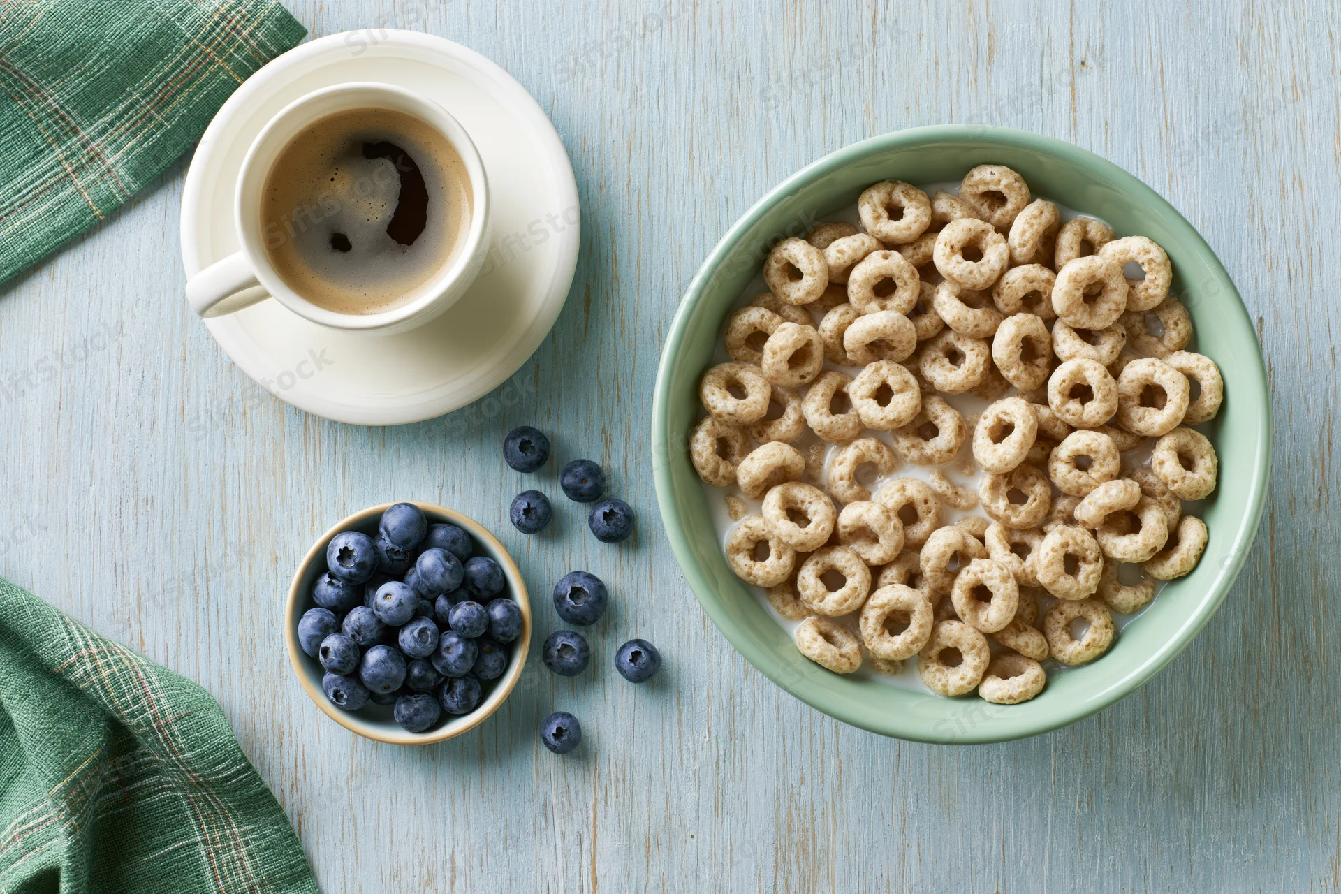 Overhead flat lay of a green bowl filled with cereal and milk, blueberries in a small bowl, coffee cup on saucer, on a light wooden table with a green plaid napkin