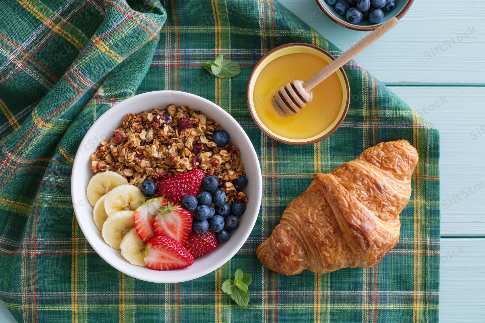 Overhead flat lay of a white bowl of granola with banana, strawberry, blueberry, and mint, next to a croissant and honey on a plaid napkin on a wooden table