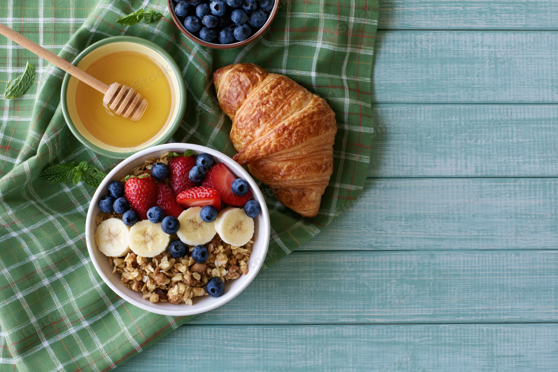 Overhead flat lay of a bowl of granola topped with banana, blueberries, strawberries and mint next to a croissant and honey on a green plaid napkin on a teal wooden table