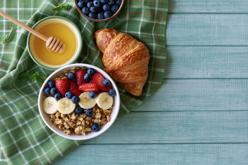 Overhead flat lay of a bowl of granola topped with banana, blueberries, strawberries and mint next to a croissant and honey on a green plaid napkin on a teal wooden table
