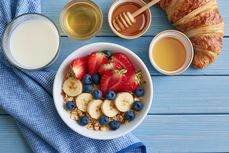 Overhead flat lay of a white ceramic bowl filled with granola, strawberries, blueberries and banana slices on a blue wooden table with milk, honey, croissant and checkered napkin