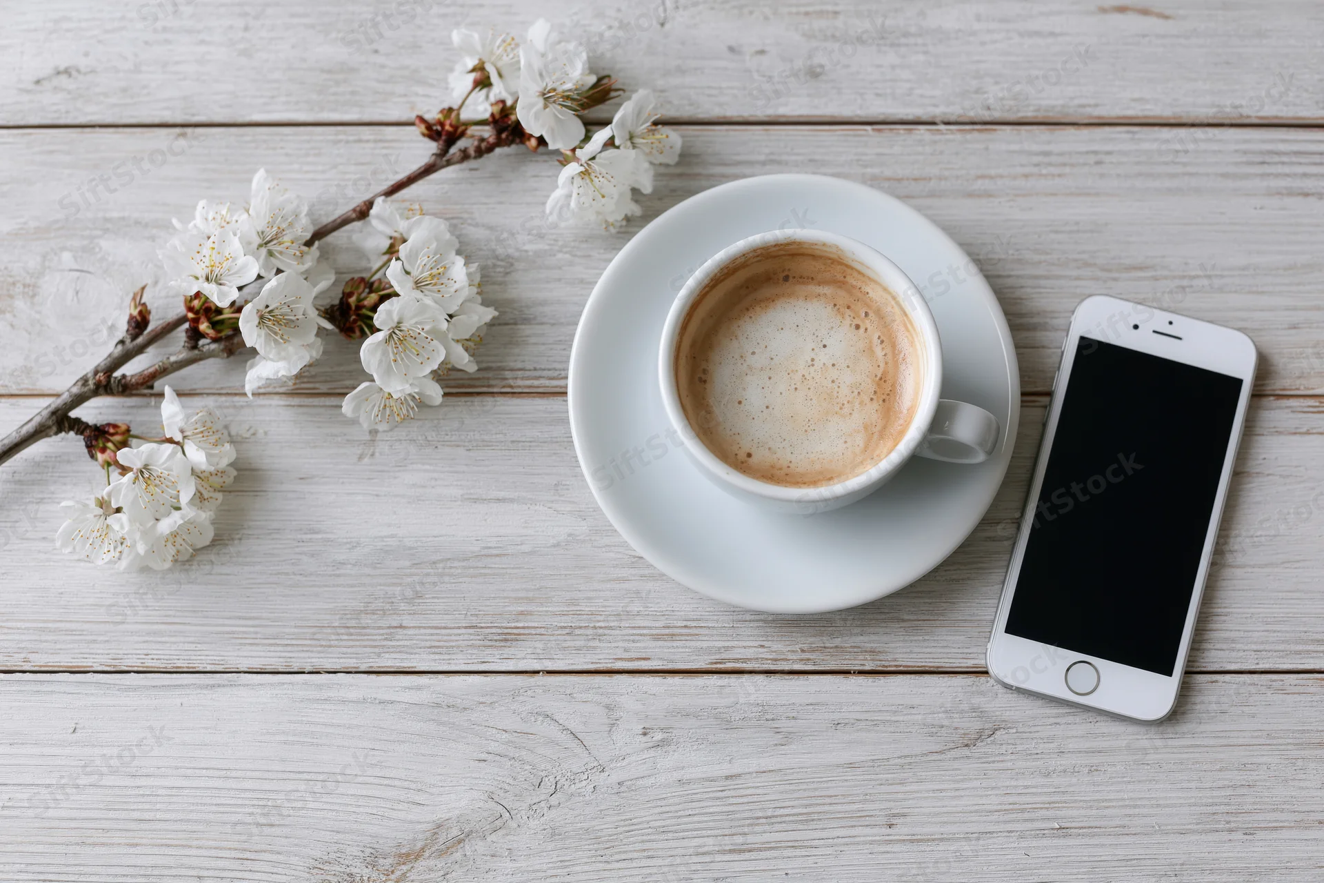 White ceramic cappuccino cup with saucer and smartphone on whitewashed wooden table beside cherry blossom branch