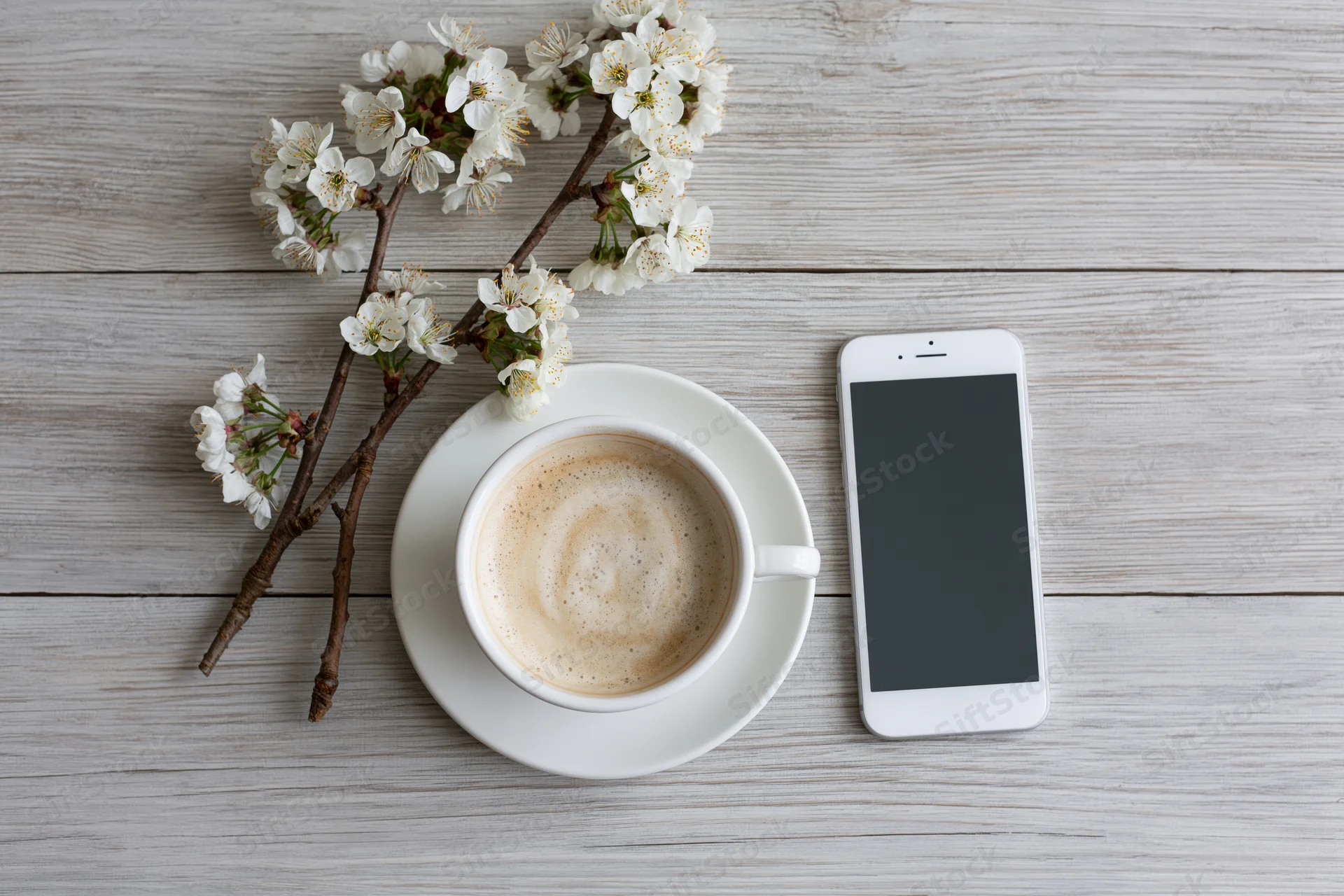 White ceramic cappuccino cup with saucer, smartphone and cherry blossom branch on gray wooden table in flat lay overhead view