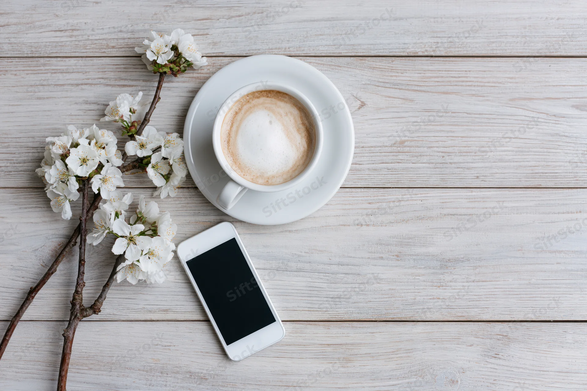 White ceramic cappuccino cup with saucer, smartphone and blooming branch on light wooden table in flat lay overhead view