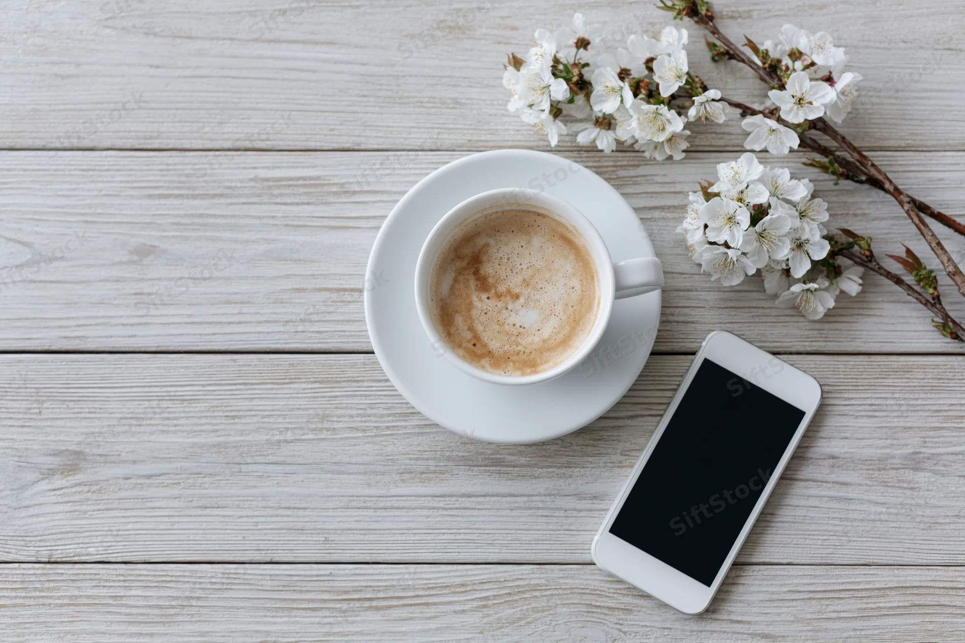 White ceramic cappuccino cup with smartphone and cherry blossoms on light wooden table in flat lay arrangement