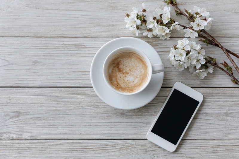 White ceramic cappuccino cup with smartphone and cherry blossoms on light wooden table in flat lay arrangement
