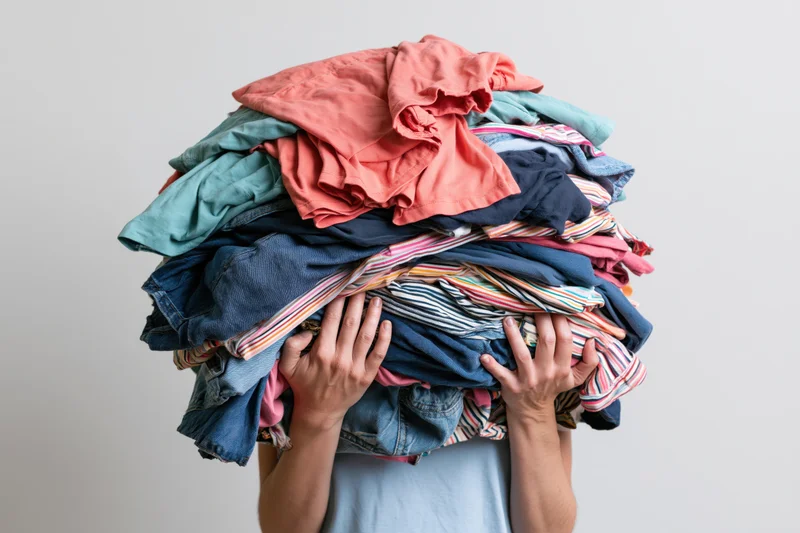 Woman holding large messy pile of clothes over her head in a plain indoor setting