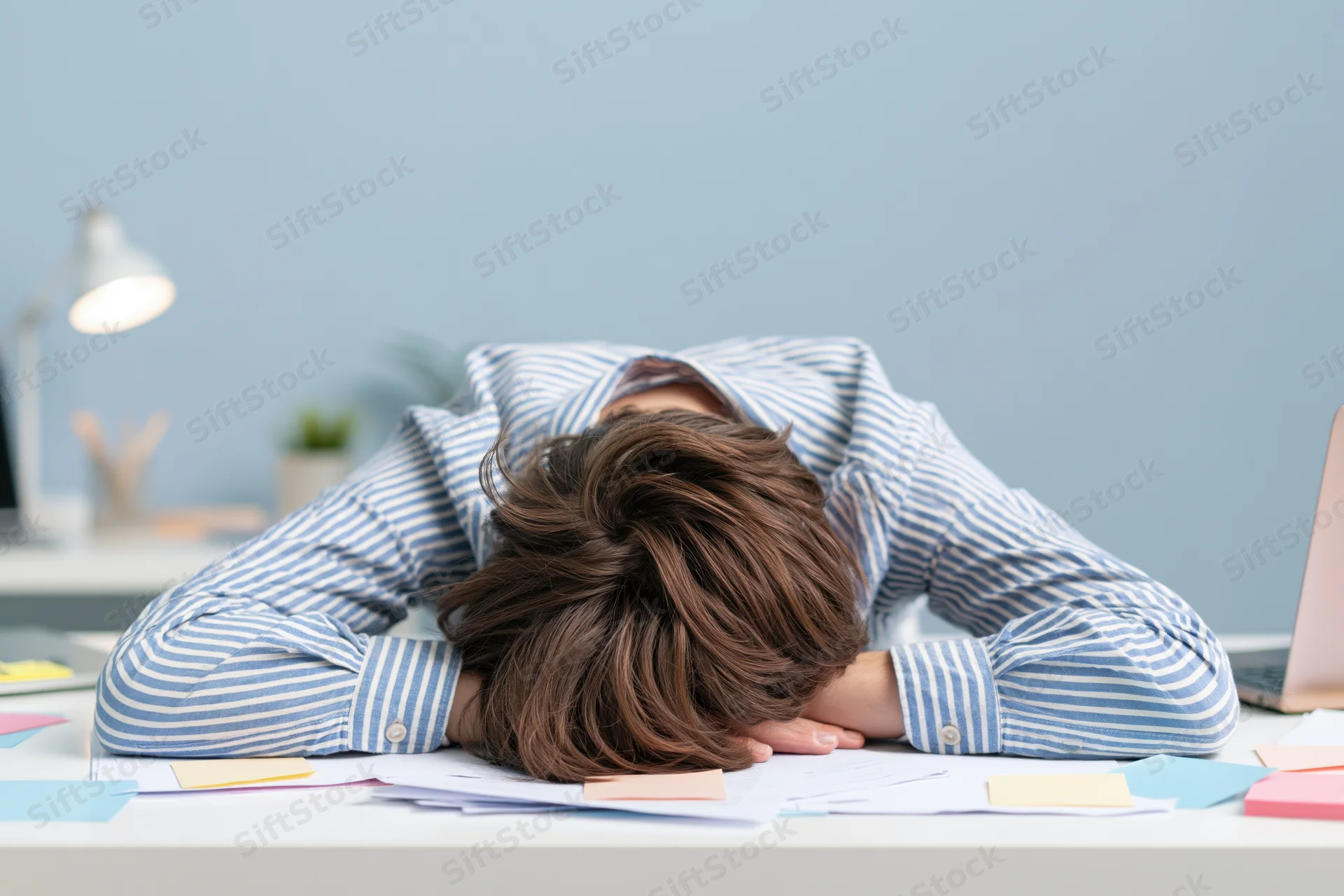 Woman slumped face-down on office desk surrounded by papers and laptop